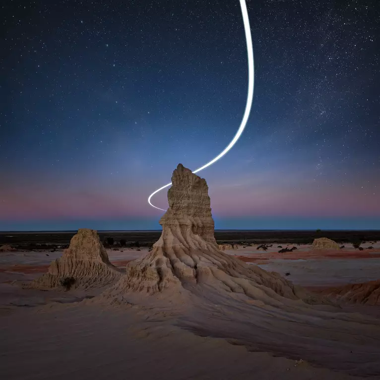 Eroded clay pinnacle in a desert landscape at dusk with a curving line of light falling from the starry sky around it