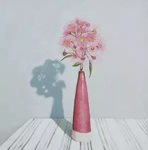 A single stem of pink flowering gum in a tall slender cranberry coloured ceramic vase on a white tablecloth with black strip…
