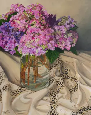 A glass vase of pink and purple hued hydrangeas placed on a draped cream antique Battenberg Lace tablecloth